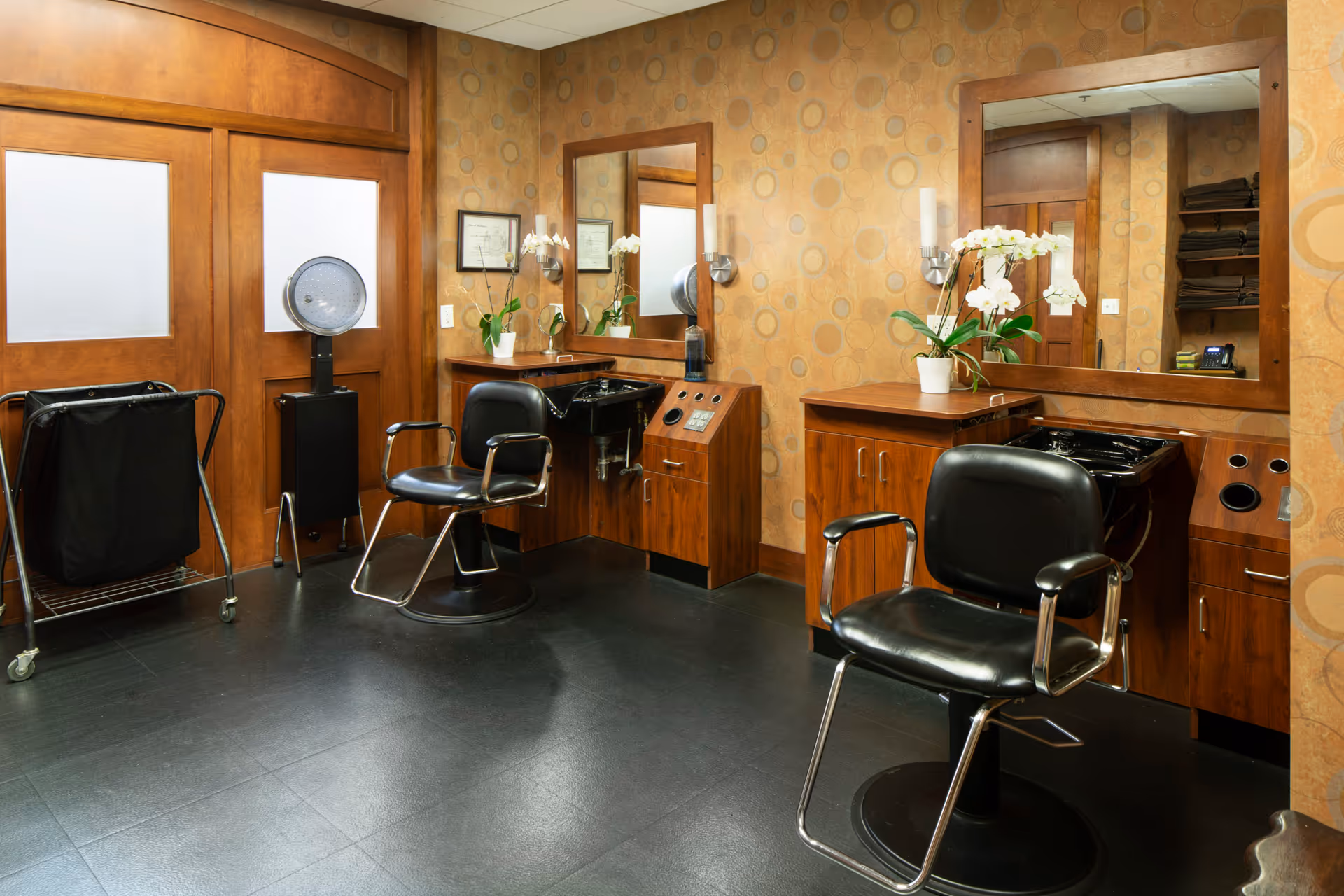 Small salon room with two black styling chairs, washbasins, mirrors, wooden cabinets, and patterned wallpaper.