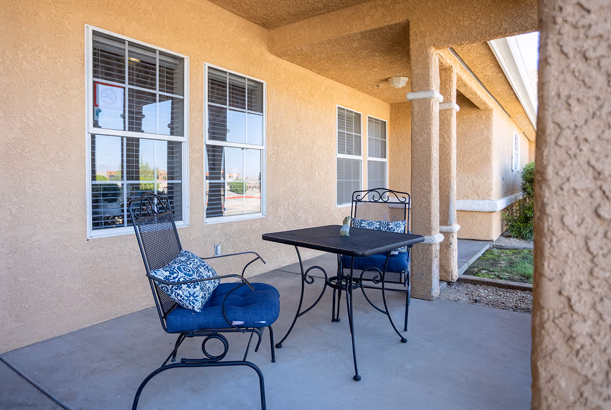 Outdoor covered patio area with two black metal chairs with blue cushions and a matching black metal table. The patio is adjacent to a beige stucco building with several windows and decorative columns.