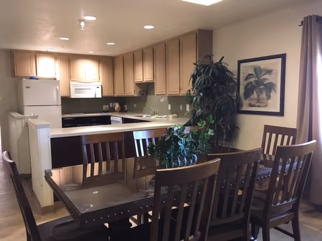 A dining area with a dark wooden table and six matching chairs. Behind the dining table is a kitchen with beige cabinets, a white refrigerator, microwave, and dishwasher. There is a large green plant next to the dining table and a framed picture of palm trees on the wall. A window with brown curtains is visible on the right side.
