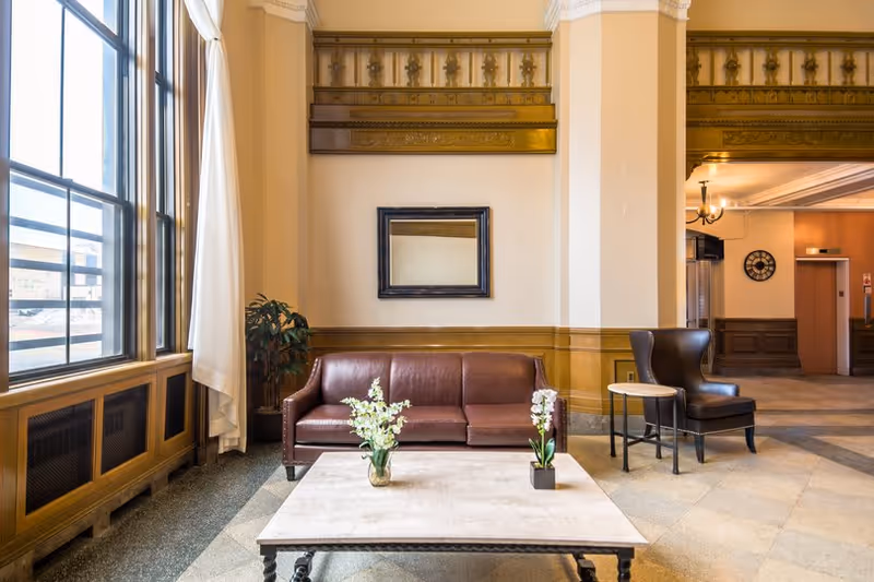 A well-lit interior lounge area with a brown leather sofa, a dark brown armchair, and a light-colored coffee table with two flower vases. Large windows with white curtains allow natural light to fill the space. The walls feature decorative molding and a framed mirror.