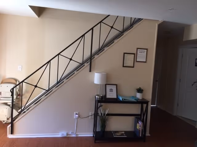 Interior view of a hallway area with a staircase featuring a black metal railing. Below the staircase is a small black console table with a white lamp, framed pictures, a small plant, and some folders. The wall behind the table has two framed documents. The floor is wooden, and there is a hallway leading to other rooms.