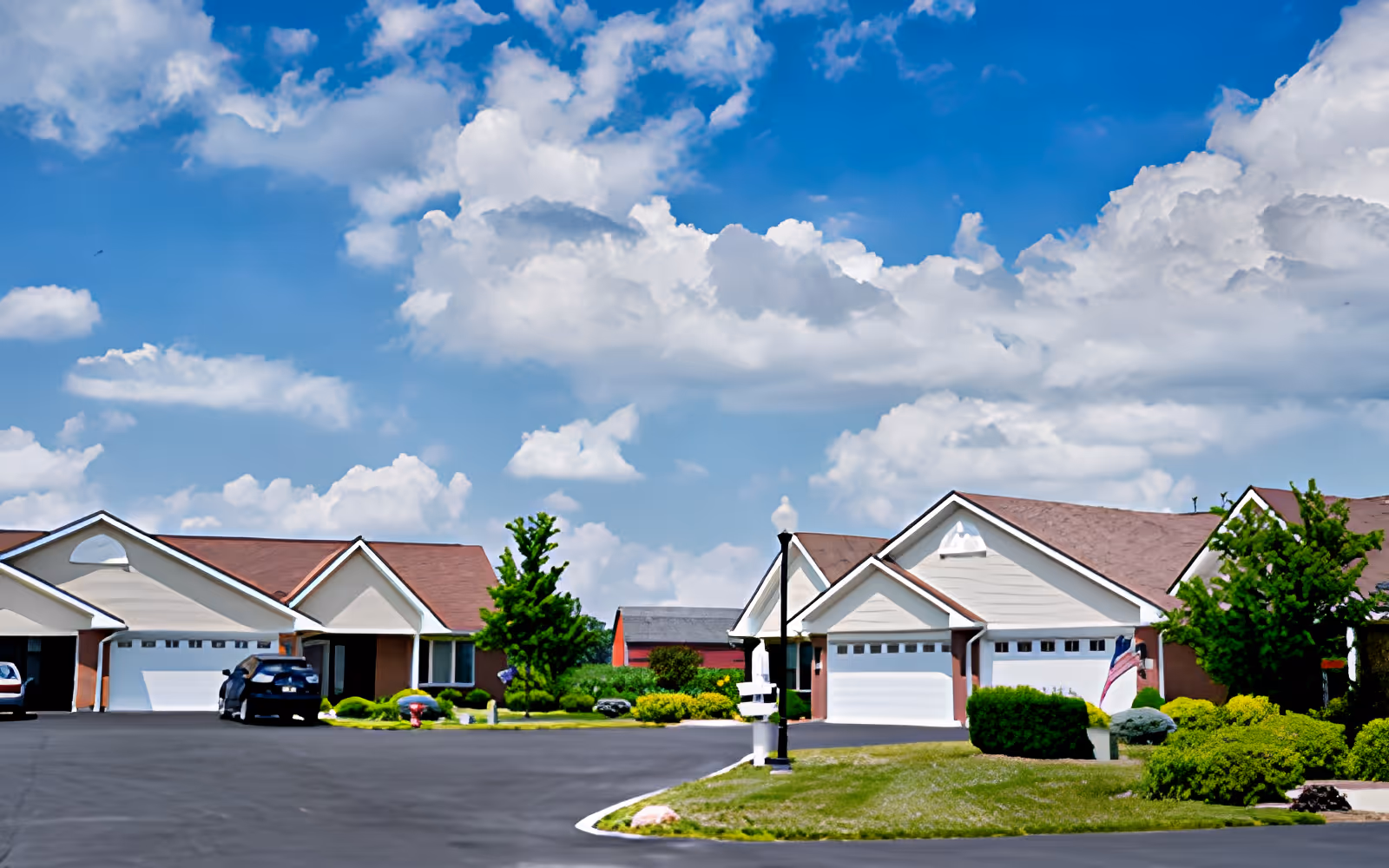 View of a quiet residential street in a retirement community with single-story houses featuring garages, well-maintained lawns, bushes, and trees under a partly cloudy blue sky.