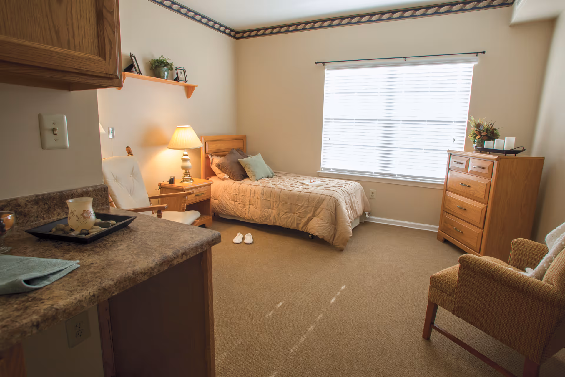 A cozy bedroom in a senior living facility with a single bed covered in a beige quilt, two pillows, and a pair of white slippers on the carpeted floor. There is a wooden nightstand with a lamp next to the bed, a wooden dresser with decorative items on top, a cushioned armchair, and a countertop with a candle and tray. A large window with blinds allows natural light into the room.