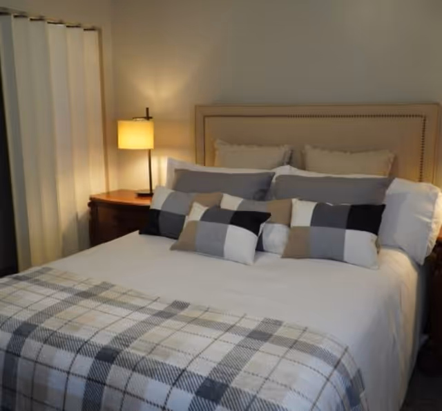 A neatly made bed with a beige upholstered headboard, multiple pillows in shades of gray, white, black, and beige, and a plaid blanket at the foot of the bed. A wooden nightstand with a lit lamp is visible on the left side of the bed, and a white curtain is partially visible in the background.