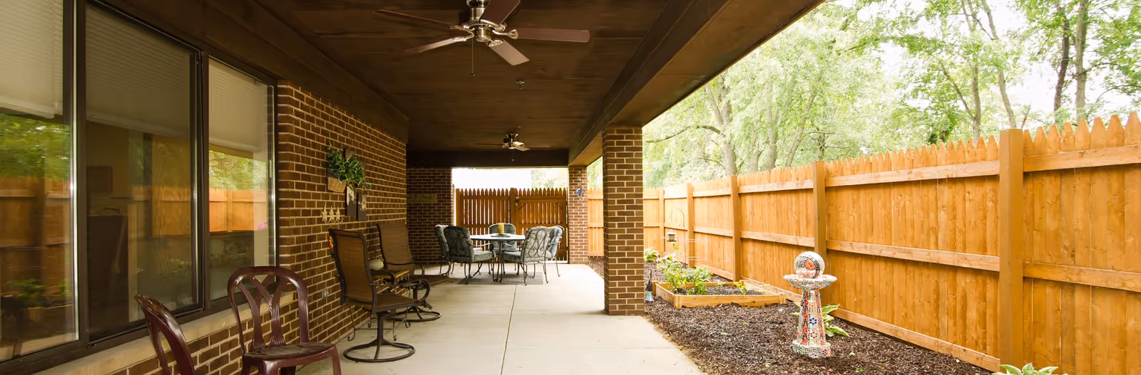 Covered outdoor patio area with ceiling fans, several chairs, a table, and a wooden fence enclosing a garden bed with plants and a decorative birdbath.