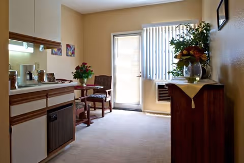 A small, cozy room with a kitchenette on the left, a round table with two chairs in the corner, and a wooden cabinet with a vase of flowers on the right. A door with vertical blinds lets in natural light, and the walls are painted beige.