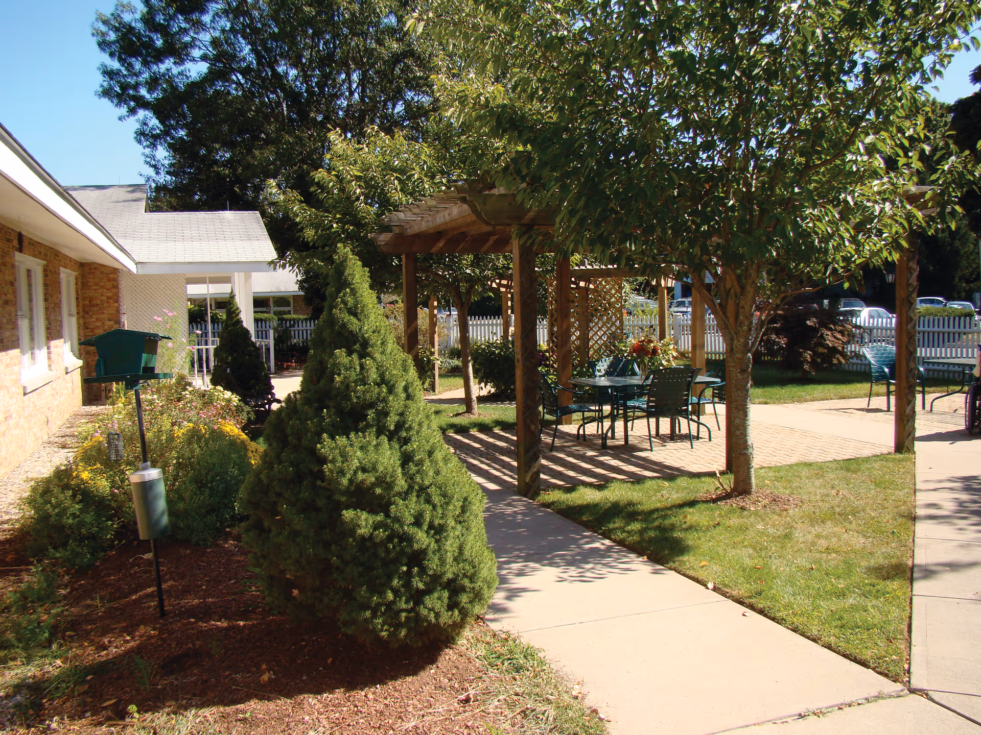 Outdoor garden area at a senior living facility with a paved walkway, green shrubs, trees, and a wooden pergola covering a patio table and chairs. A brick building is visible on the left side, and a white picket fence encloses the area.