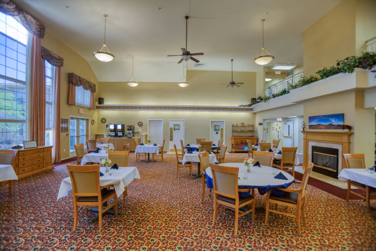 Spacious dining room with multiple round and square tables covered with white tablecloths and set with napkins and small flower arrangements. The room features large windows with curtains, a patterned carpet, ceiling fans, pendant lights, a fireplace with a landscape painting above it, and a beverage station in the back.