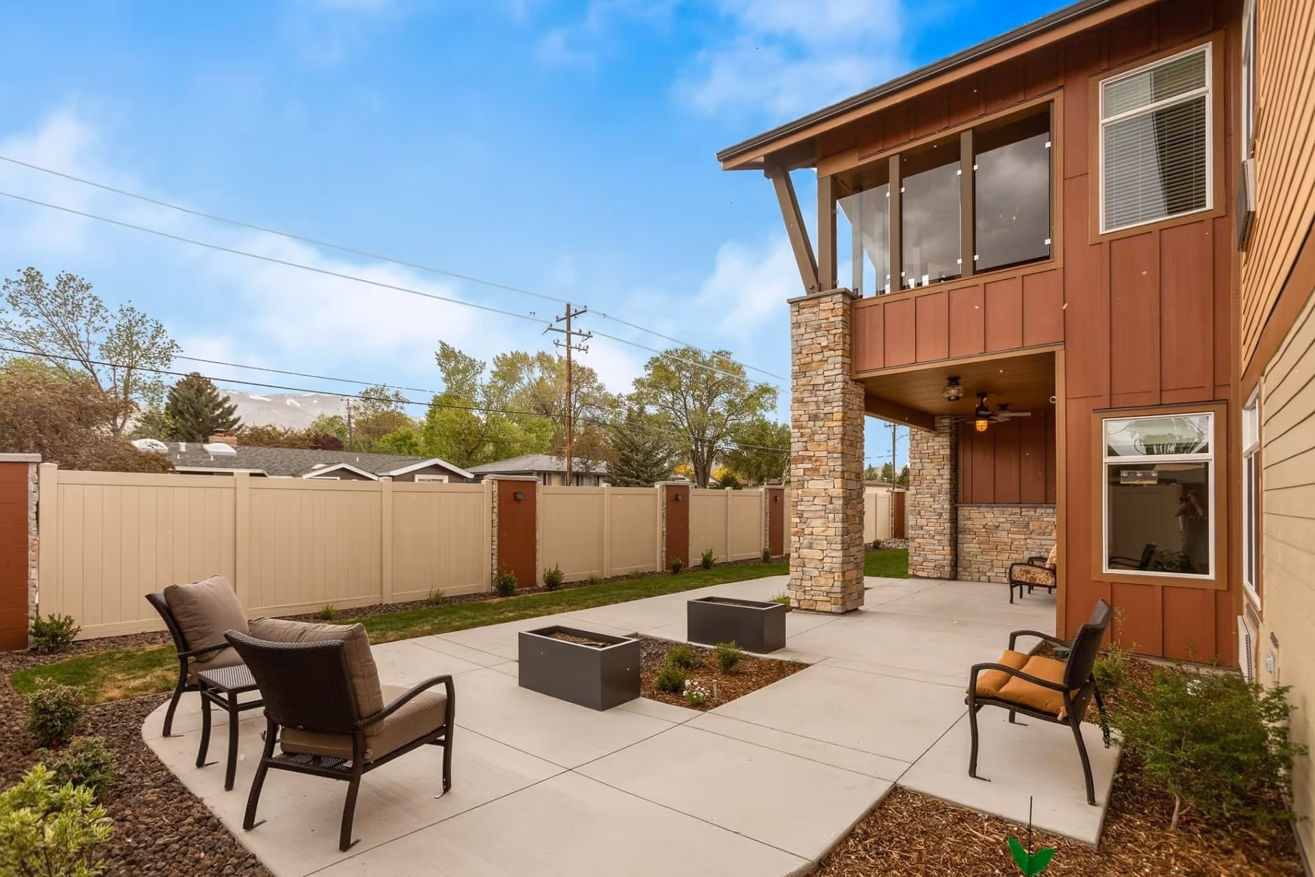 Outdoor patio area at Sierra Basin Memory Care with cushioned chairs arranged around two square planters on a concrete surface, surrounded by a beige privacy fence and landscaping with small plants and trees under a clear blue sky.