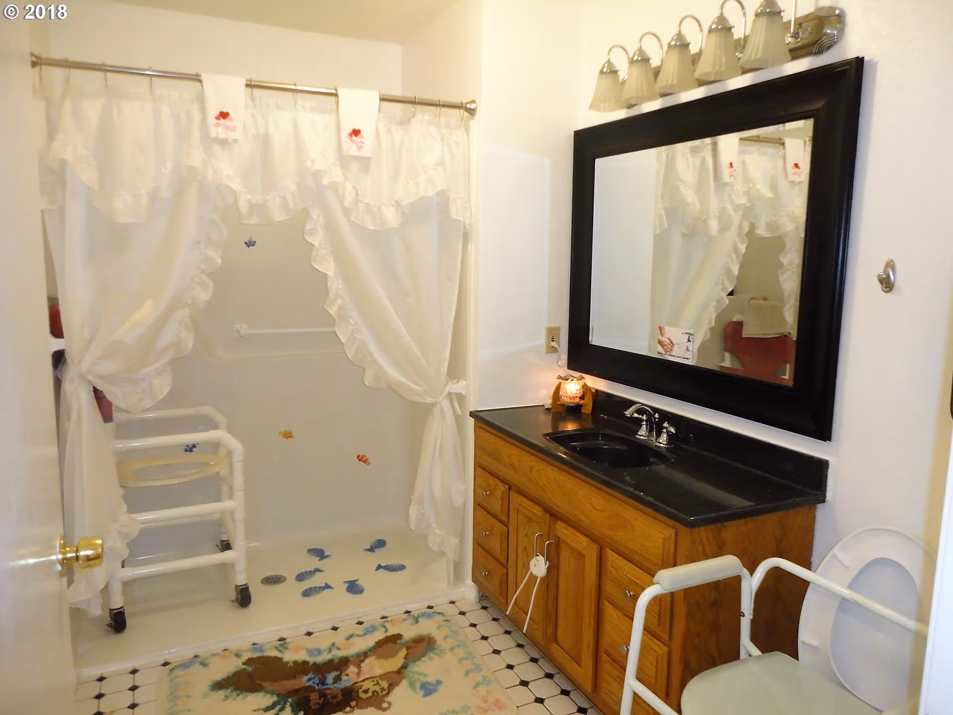 Bathroom with a walk-in shower featuring white ruffled curtains, a white shower chair, and fish decals on the shower floor and wall. There is a wooden vanity with a black countertop and sink, a large black-framed mirror above it, and a light fixture with five lights. A toilet with safety rails is visible on the right side, and a floral-patterned rug is on the tiled floor.