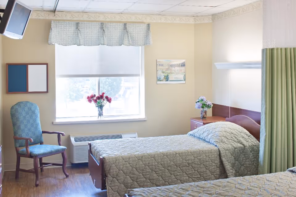 A bright and clean bedroom with two single beds covered in light gray quilted bedspreads. There is a wooden nightstand between the beds with a vase of flowers on it. A window with a light-colored valance and a roller shade lets in natural light. A blue upholstered chair is positioned near the window, and a wall-mounted TV is visible in the upper left corner. The room has light-colored walls and wood flooring.