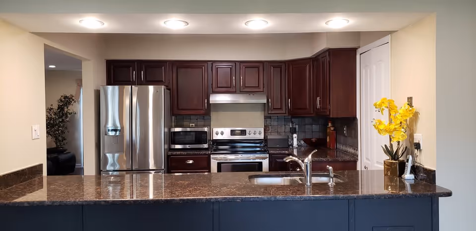 Modern kitchen with dark wooden cabinets, stainless steel refrigerator, microwave, stove, and a sink on a granite countertop. A yellow orchid plant in a gold pot is placed on the right side of the counter.