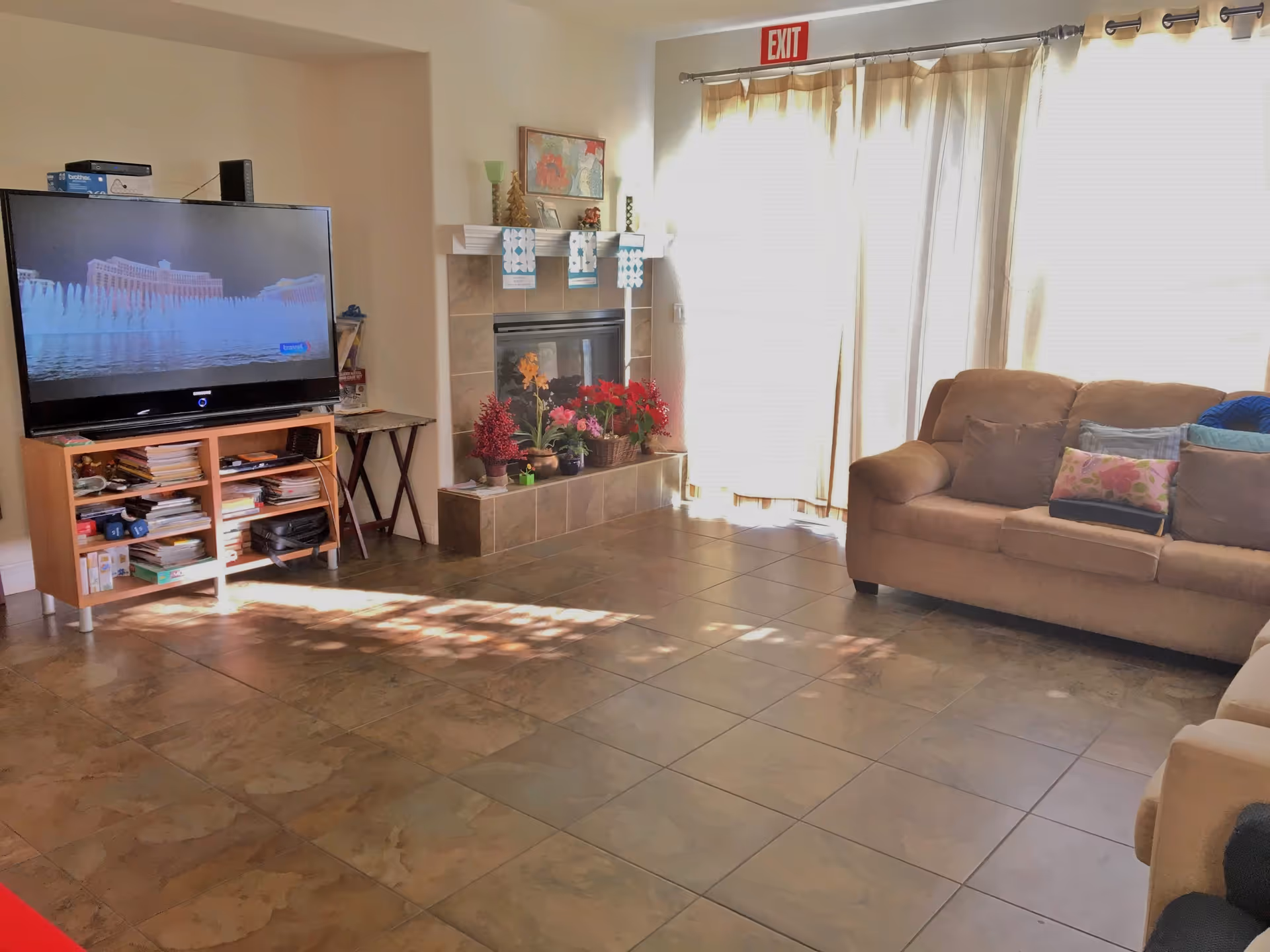 A cozy living room with a large flat-screen TV on a wooden stand filled with books and other items. There is a fireplace decorated with plants and holiday ornaments. A beige sofa with multiple cushions is positioned near a large window covered with light curtains, and an exit sign is visible above the window.