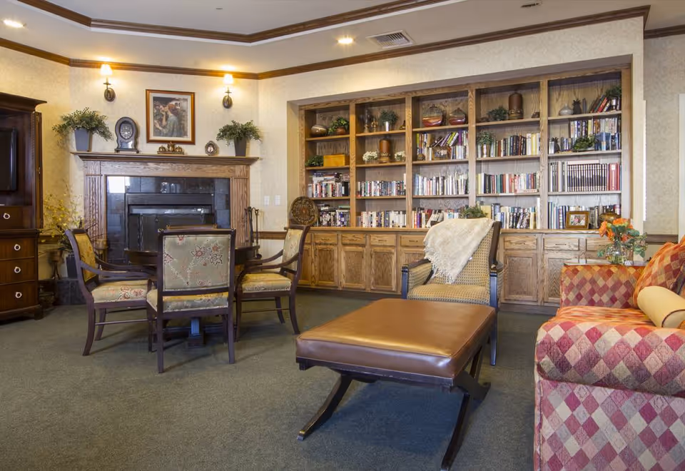 A cozy living room area with a fireplace, wooden built-in bookshelves filled with books and decorative items, a round table with four upholstered chairs, a cushioned bench, and a patterned sofa with a yellow bolster pillow. The room has warm lighting and a carpeted floor.