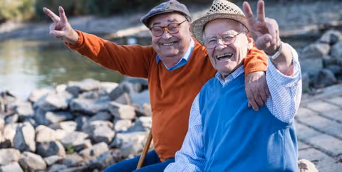 Two elderly men sitting outdoors on a stone embankment near a body of water, smiling and making peace signs with their hands. One man is wearing an orange sweater and a cap, and the other is wearing a blue sweater vest and a straw hat.