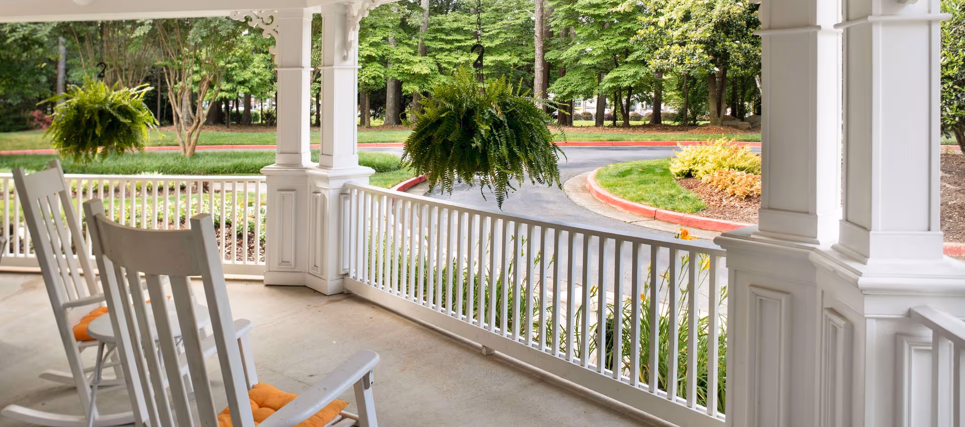 Front porch with white rocking chairs and hanging ferns overlooking a curved driveway and landscaped grounds.