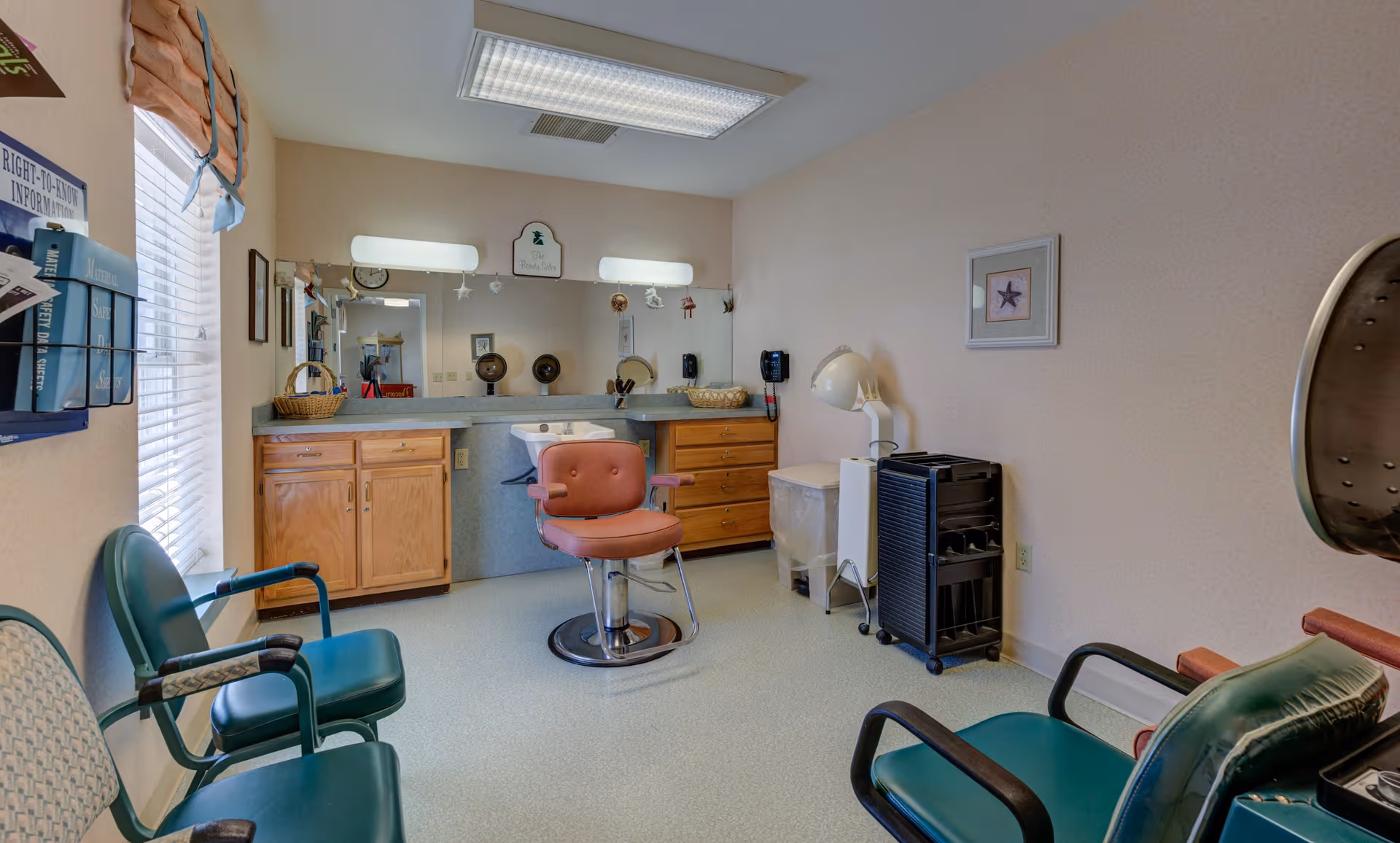 Interior of a senior living community hair salon with a pink salon chair in front of a large mirror, wooden cabinets, green waiting chairs, a hair dryer, and various salon equipment.