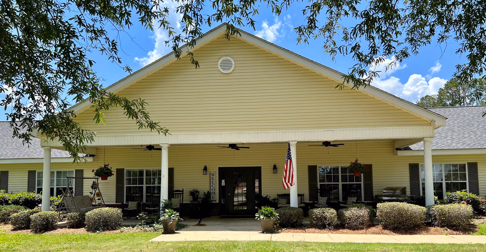 Front exterior view of a single-story yellow building with a covered porch supported by white columns. The porch has hanging flower pots, rocking chairs, an American flag, and a welcome sign near the entrance. The building is surrounded by neatly trimmed bushes and trees, under a partly cloudy blue sky.