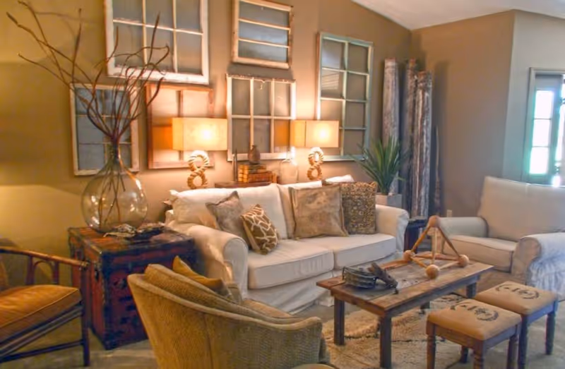 A cozy living room with a white sofa adorned with multiple patterned cushions, a wooden coffee table with decorative items, two cushioned stools, and additional seating including a chair and an armchair. The wall behind the sofa features vintage window frames and two table lamps emitting warm light. A large glass vase with branches and a potted plant add natural elements to the room.