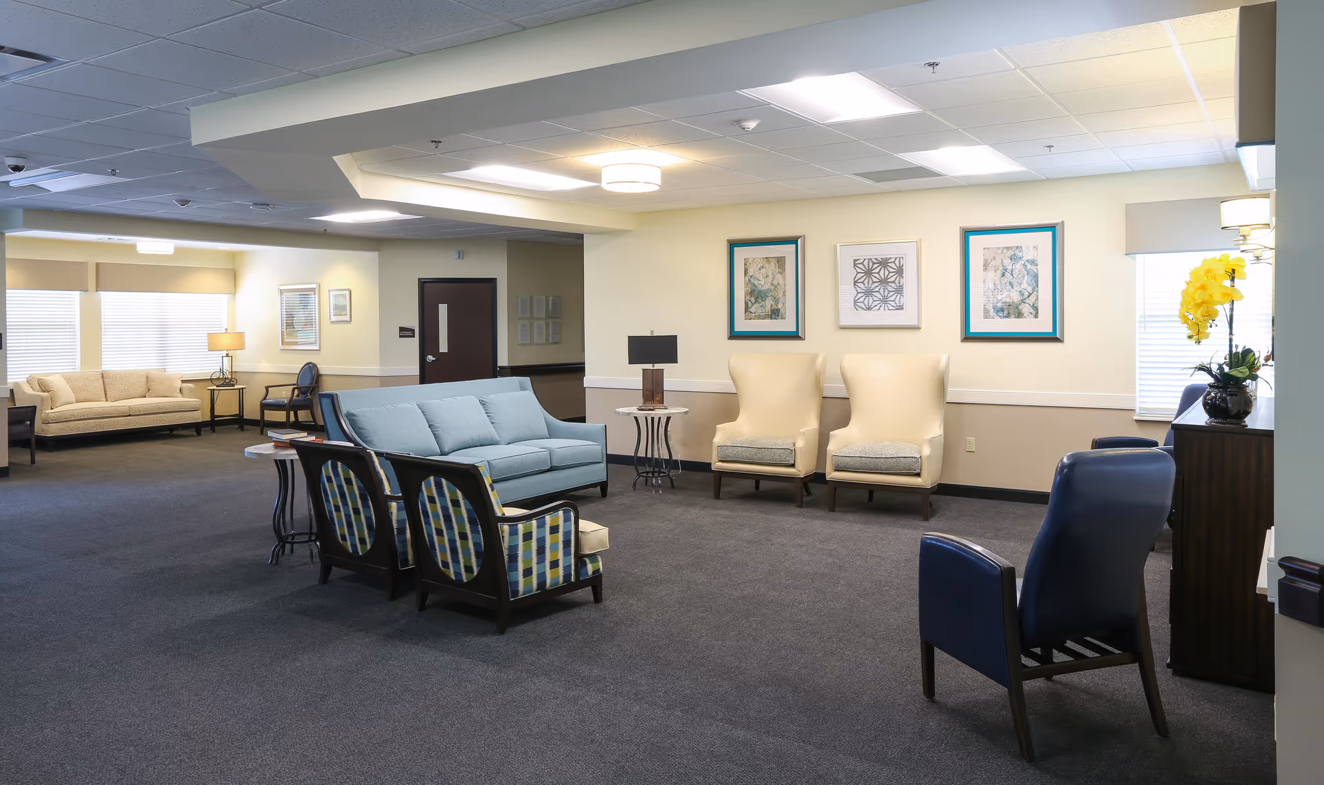 A spacious senior living facility common area with various seating options including a light blue sofa, two patterned armchairs, two cream-colored armchairs, and two dark blue chairs. The room has neutral walls with framed artwork, carpeted flooring, and large windows letting in natural light. There are side tables with lamps and a cabinet with a yellow orchid plant.