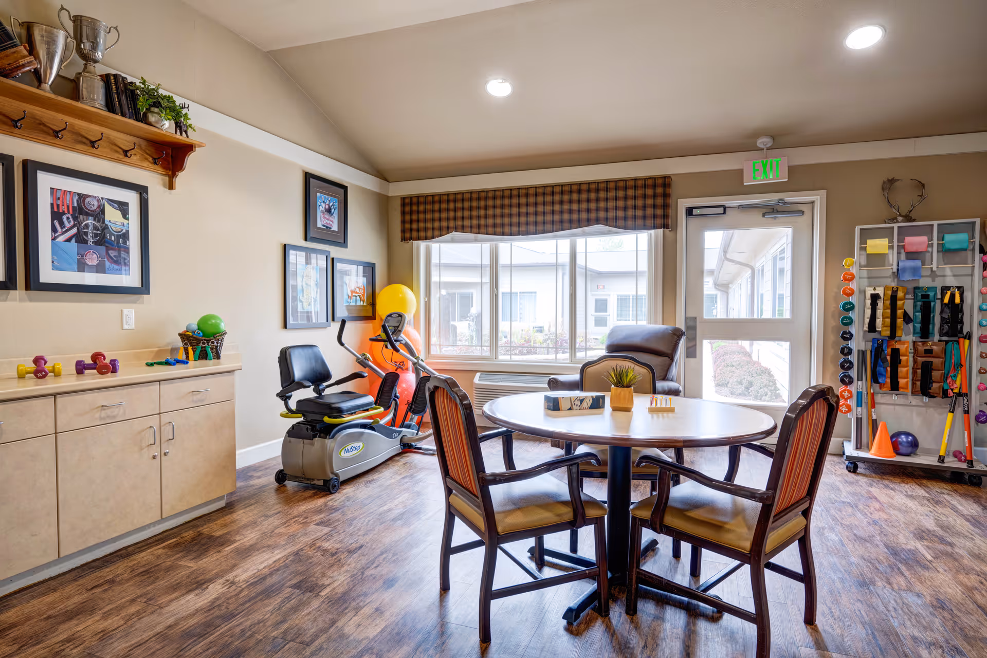 A bright room with wooden flooring featuring a round table with four chairs, a comfortable recliner chair near a large window, and exercise equipment including a stationary bike and various weights and resistance bands on a rack. The room has beige walls with framed pictures and a shelf with trophies and plants.