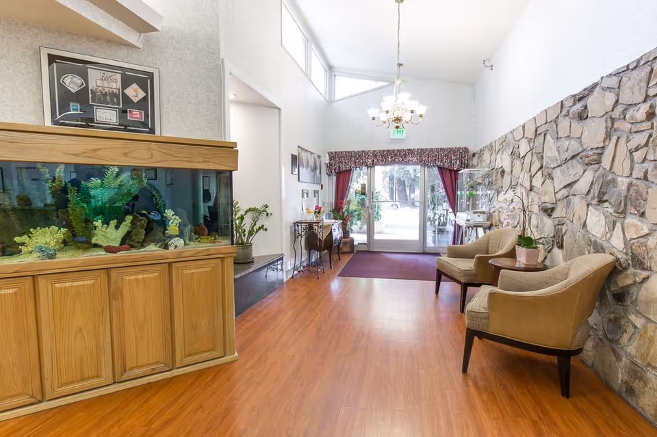 Entrance lobby with a large aquarium on the left, two chairs and a stone accent wall on the right, and glass doors under a chandelier leading outside.