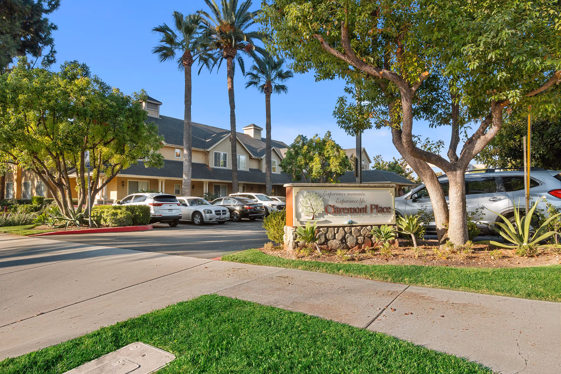 Exterior view of Claremont Place senior living facility with a parking lot, several cars, palm trees, and other greenery under a clear blue sky. A stone sign in front reads 'Experience moments. Experience life. Claremont Place Independent Assisted Memory Care.'