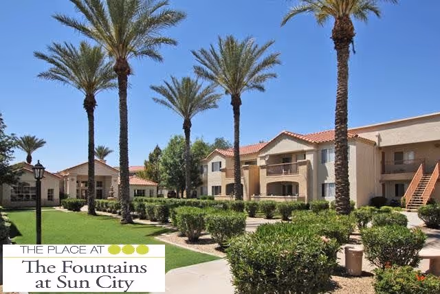 Outdoor view of a senior living facility with beige two-story buildings featuring red tile roofs, surrounded by palm trees, green bushes, and well-maintained lawns under a clear blue sky.