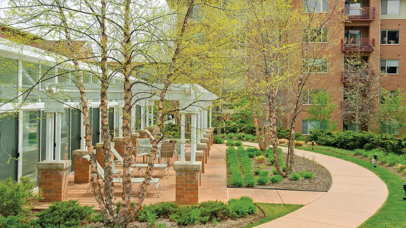 Outdoor patio area at Fox Run Senior Living Community with chairs and tables under a white pergola, surrounded by trees and landscaped greenery, with a curved walkway leading past the patio and a multi-story brick building in the background.