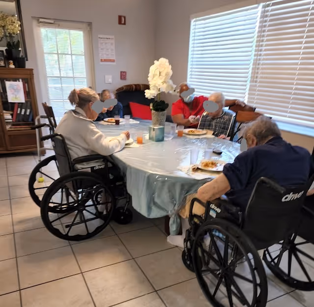 Several elderly residents in wheelchairs sit around a table in a dining area eating a meal.