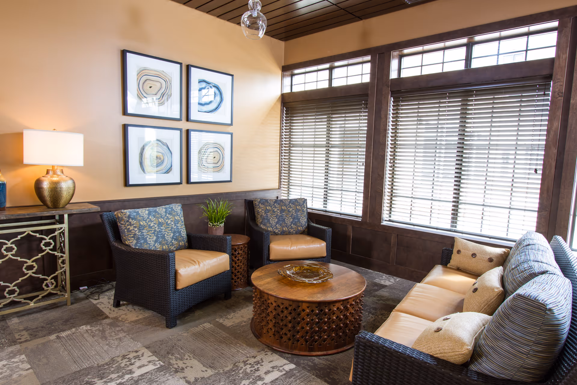 Cozy lounge with wicker armchairs and a sofa arranged around a round wooden coffee table beneath large windows with blinds.
