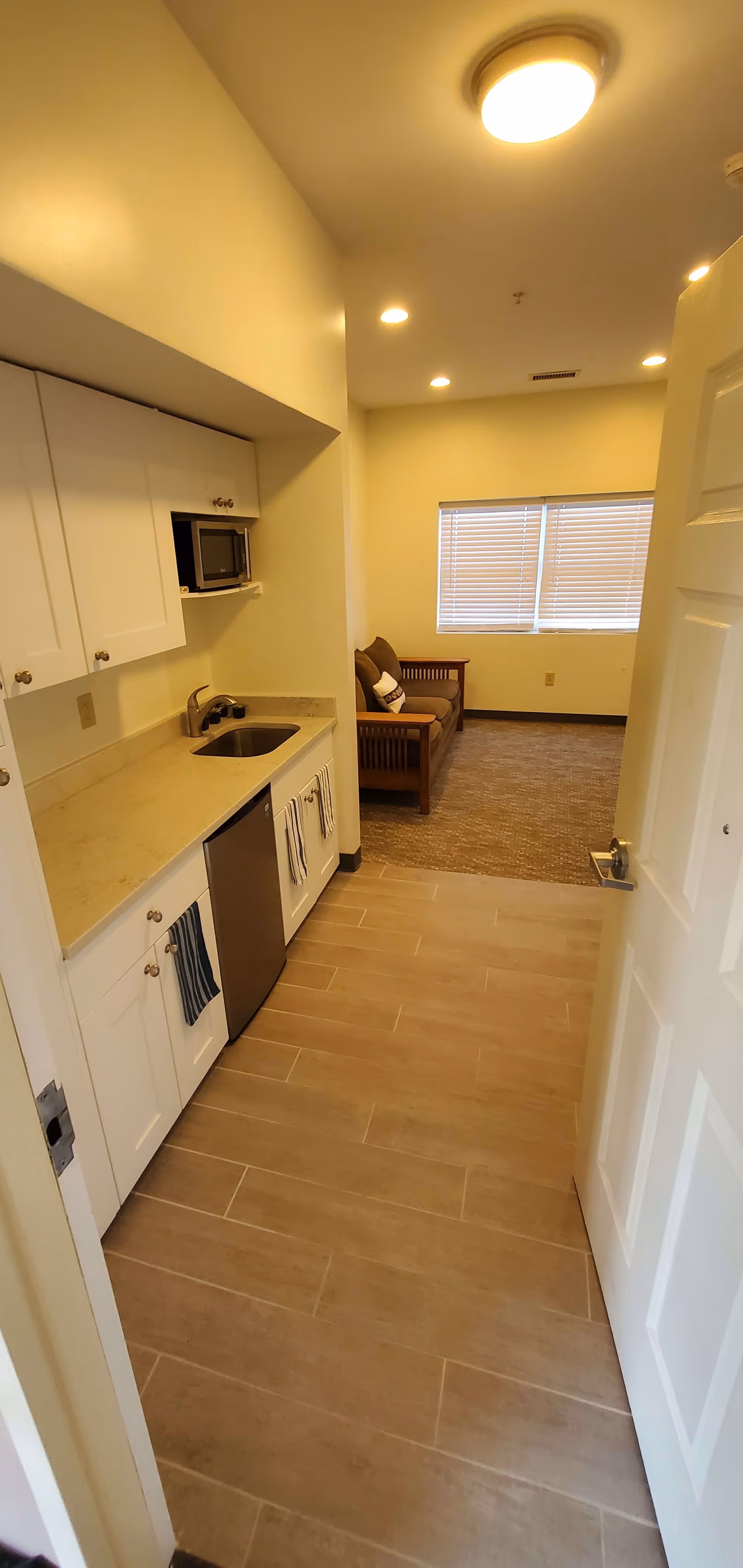 Small kitchenette with a sink, microwave and mini-fridge opening into a living area with a sofa beneath a window.