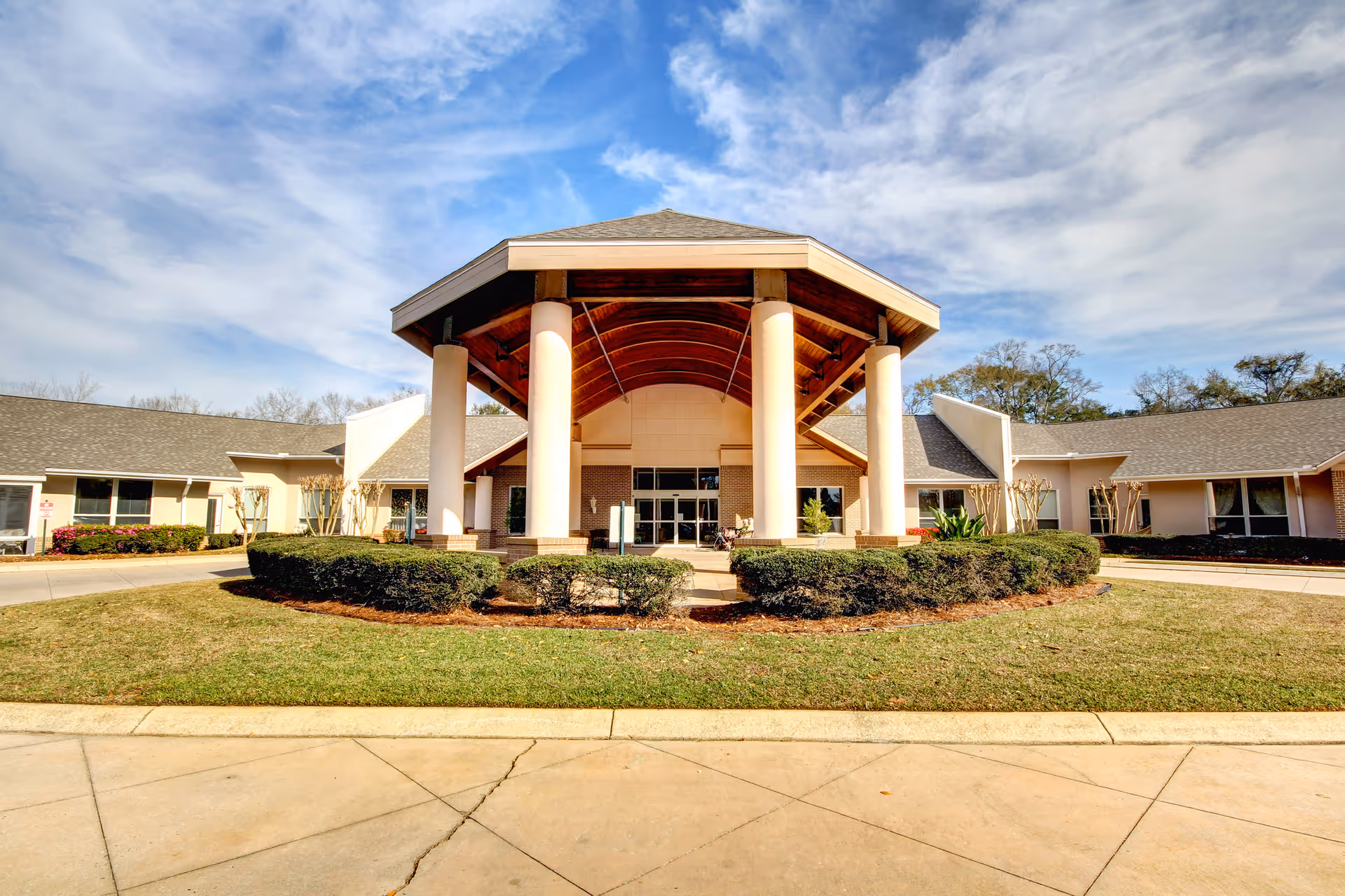 Front exterior view of Live Oak Village building with a covered entrance supported by large white columns, surrounded by trimmed bushes and a lawn under a partly cloudy sky.
