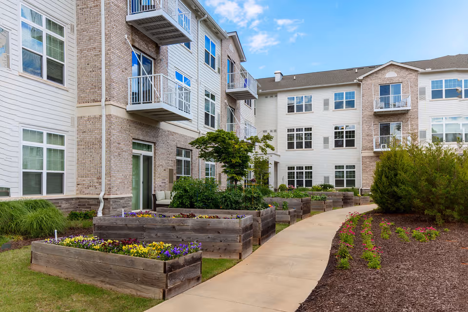 Outdoor garden area at The Sheridan at Eastside with raised wooden flower beds filled with colorful flowers, a paved walkway, and a multi-story residential building with balconies and large windows in the background under a partly cloudy sky.