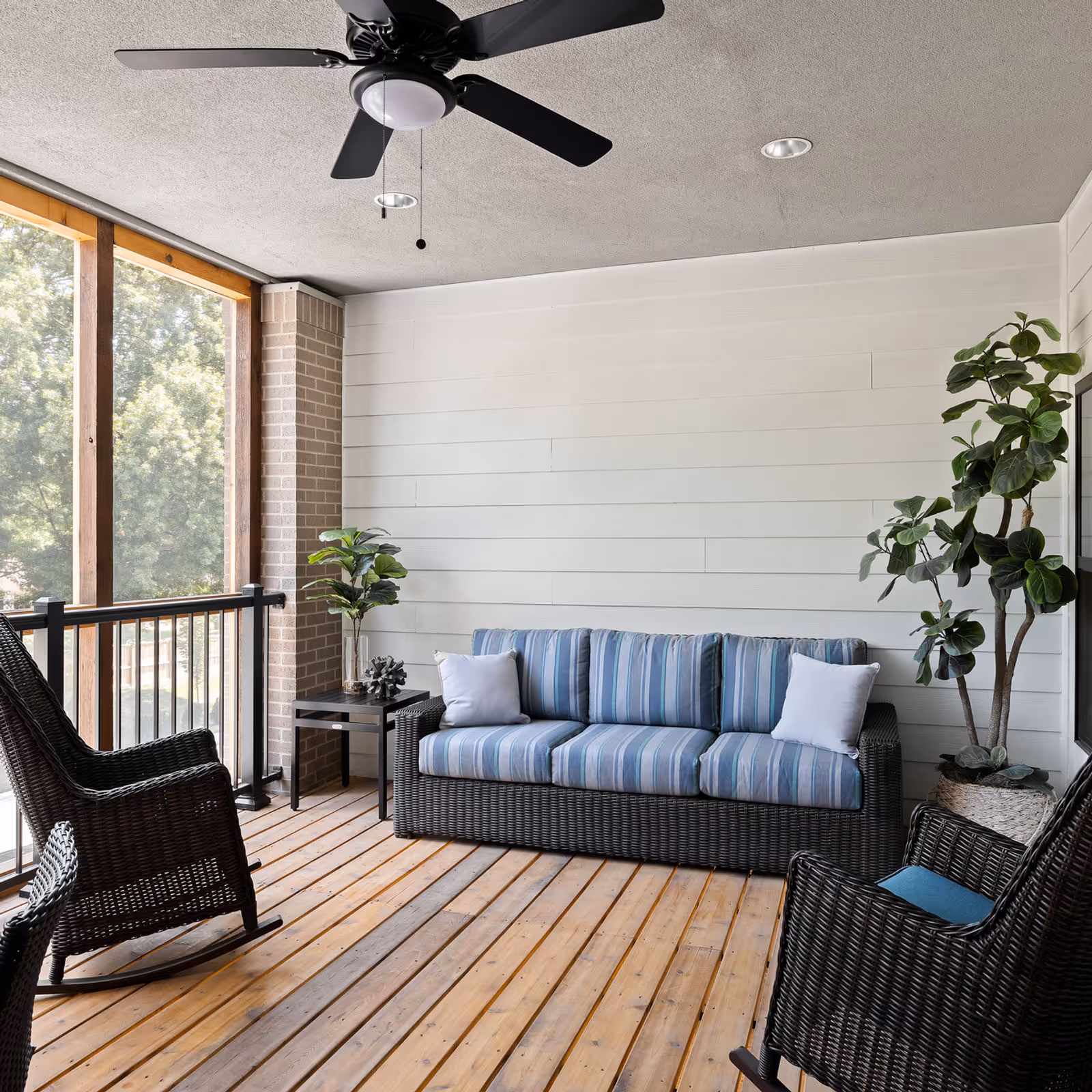 A cozy screened-in porch with wooden flooring, featuring a wicker sofa with blue striped cushions and two white pillows, two wicker rocking chairs with blue cushions, a small black side table with a decorative plant and sculpture, a large potted plant in the corner, and a ceiling fan above. The porch is enclosed with wooden framed screens allowing a view of green trees outside.