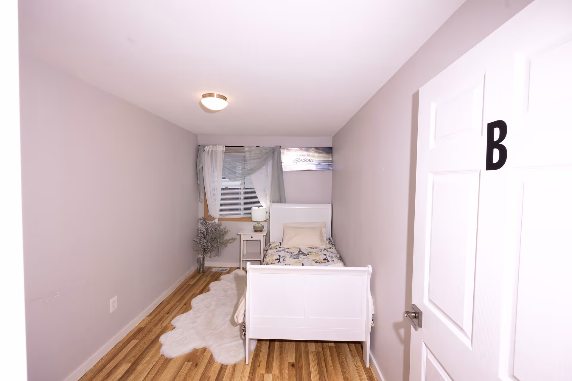 A small bedroom with light purple walls and wooden flooring. The room contains a single white bed with a floral bedspread and a beige pillow. Next to the bed is a white nightstand with a lamp and a small plant. A window with white blinds and sheer curtains is above the nightstand. A white door with the letter B is partially open on the right side of the image.