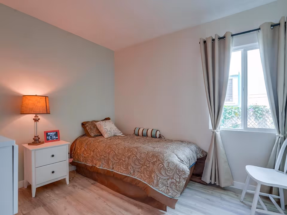 A small bedroom with a single bed covered in a brown patterned comforter and pillows. Next to the bed is a white nightstand with two drawers, a lamp with a brown shade, and a digital clock displaying the time and day. There is a window with beige curtains tied back, and a white chair is placed near the window. The floor is light-colored wood.
