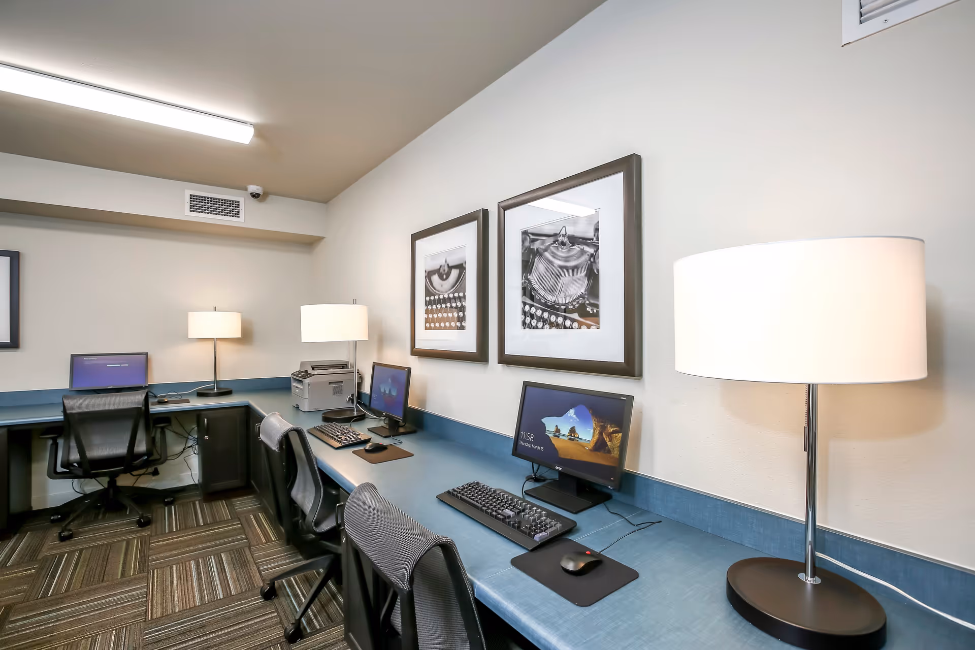 A computer room with a long blue desk holding three desktop computers, keyboards, and mice. There are three black office chairs in front of the desk. Two table lamps with white shades are placed on the desk, and two black and white framed photographs hang on the wall above the desk. The room has a patterned carpet and a ceiling light fixture.