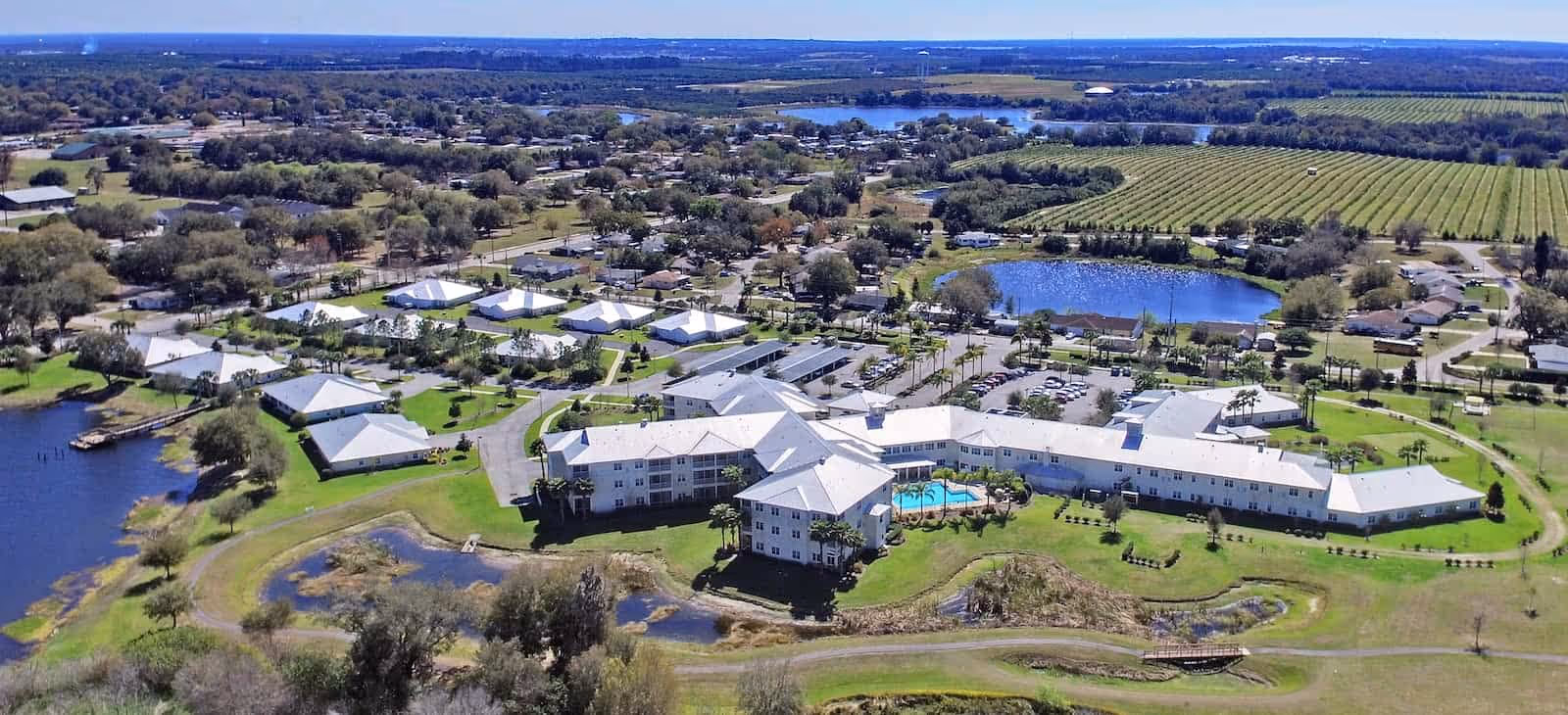 Aerial view of Water's Edge of Lake Wales senior living facility showing multiple white-roofed buildings surrounded by green lawns, trees, a swimming pool, parking lots, ponds, and nearby farmland under a clear blue sky.