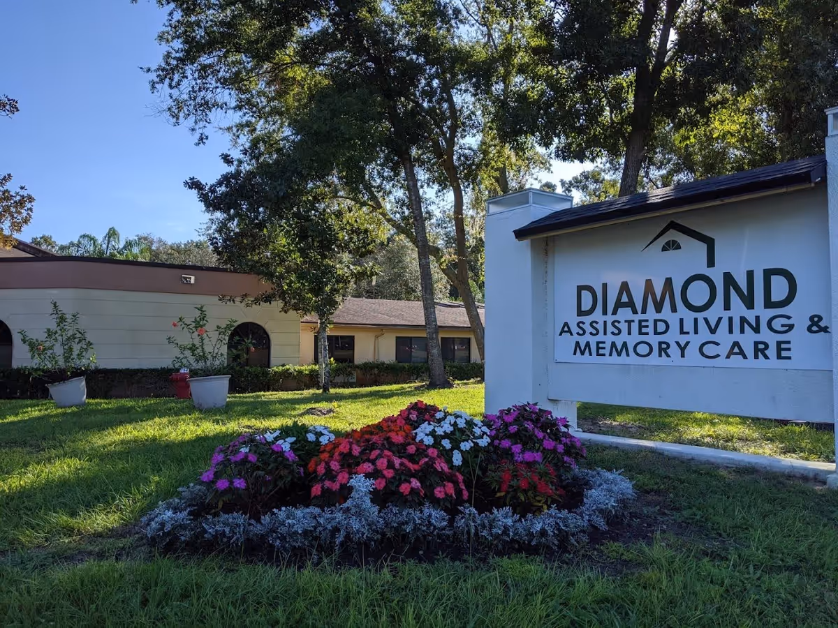 Outdoor view of Diamond Assisted Living & Memory Care facility with a sign displaying the facility's name. The scene includes a flower bed with colorful flowers, green grass, trees, and part of the building in the background under a clear blue sky.