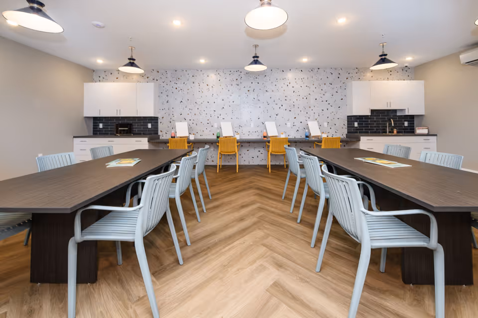 A well-lit room with two long dark wooden tables surrounded by light gray chairs. At the back wall, there are four yellow chairs placed in front of a counter with white cabinets above and below. The wall behind the counter has a terrazzo pattern, and the floor has a wood herringbone design. The ceiling has several pendant lights.