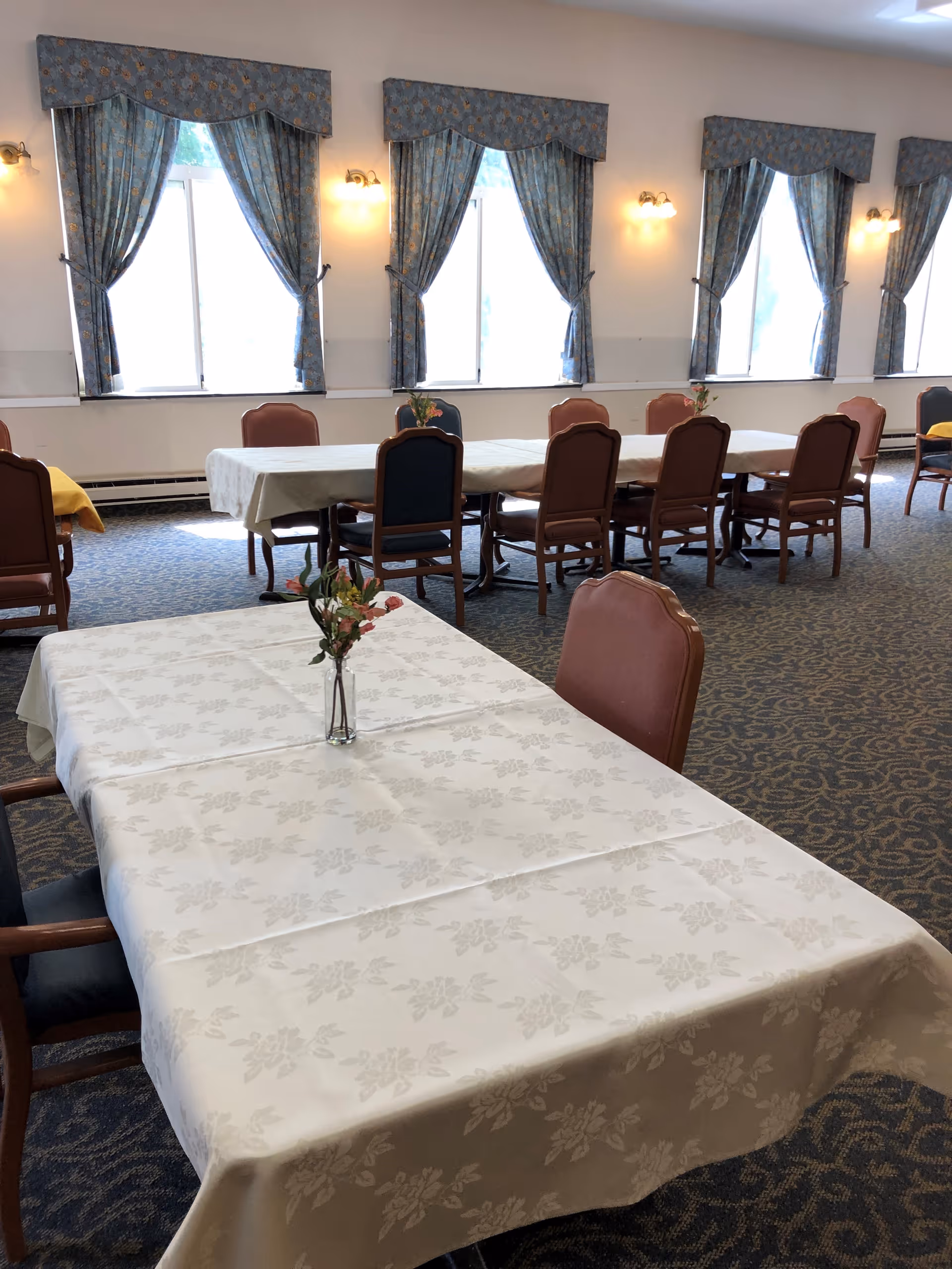 A dining room with several tables covered in white tablecloths, each with a small vase of flowers. The room has multiple windows with blue patterned curtains and wall-mounted lights between the windows. Chairs with brown and blue upholstery are arranged around the tables.