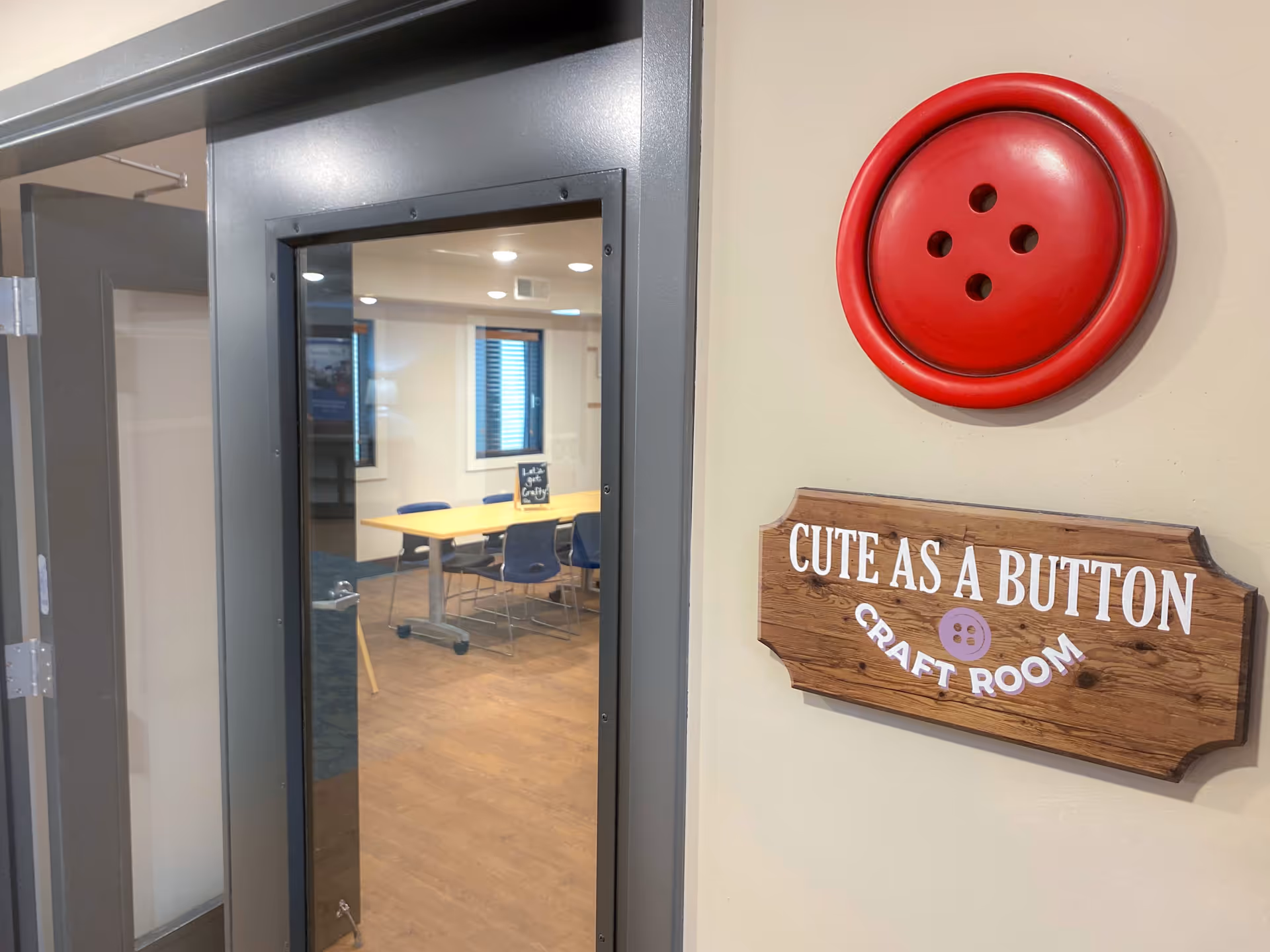 View of a craft room through a glass door with a sign on the wall next to the door that reads 'Cute As A Button Craft Room' and a large decorative red button above the sign. Inside the room, there is a table with several chairs around it and windows on the far wall.