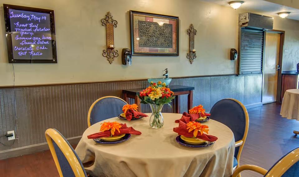 A dining room table set for four with a beige tablecloth, yellow plates, dark blue chargers, red folded napkins with orange flowers, and a vase of colorful flowers in the center. The room has wooden flooring, beige walls with decorative wall sconces, a framed artwork, and a menu board on the wall listing the meal for Tuesday, May 14. There is a hand sanitizer dispenser mounted on the wall and a serving window with a shutter in the background.
