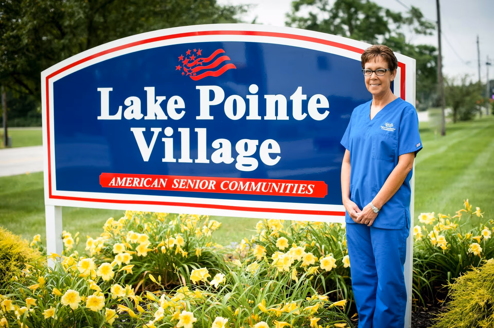 A staff member in blue scrubs stands beside a large 'Lake Pointe Village' sign surrounded by yellow flowers.