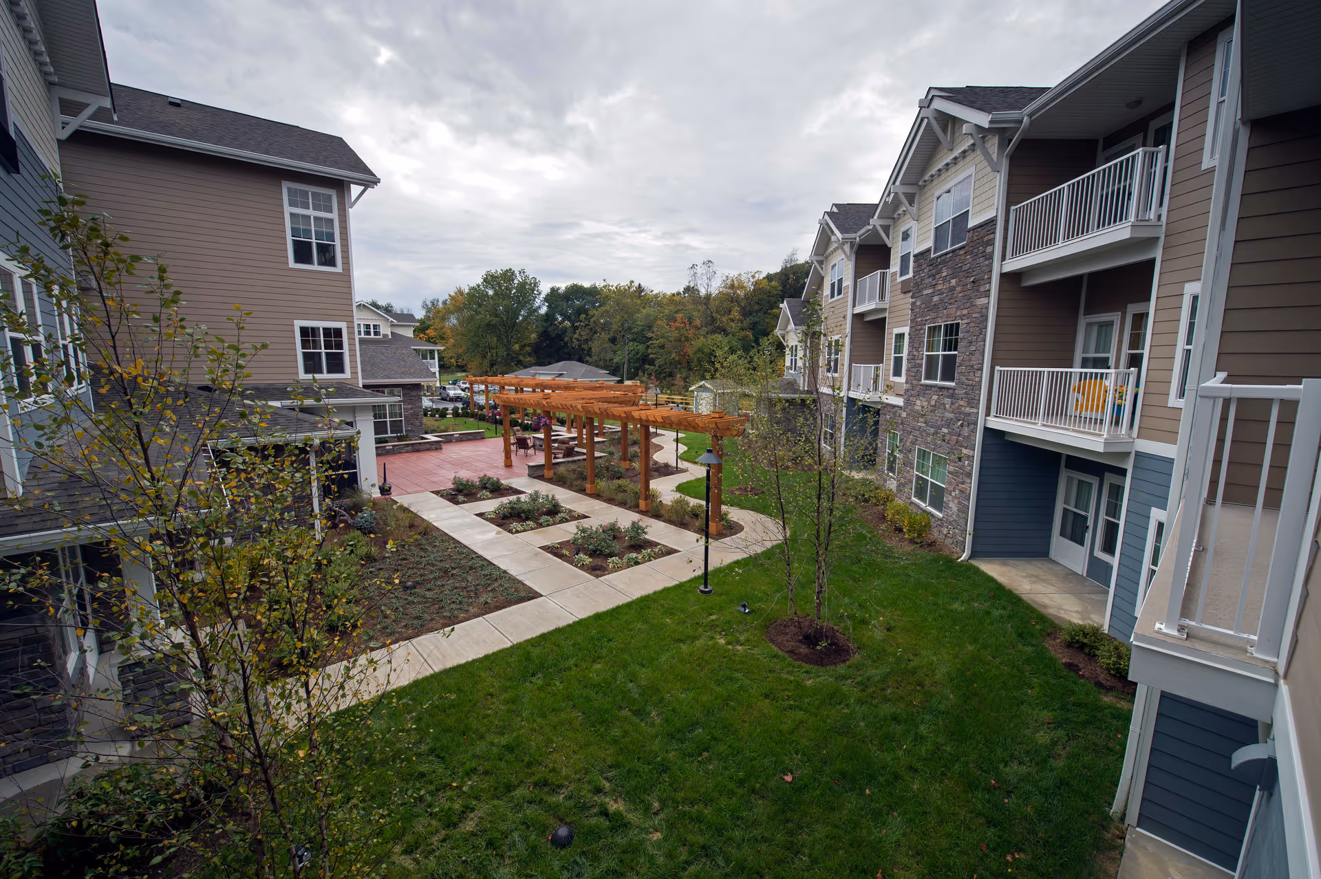 Outdoor courtyard area of a senior living facility with green grass, young trees, paved walkways, and wooden pergolas. Surrounding the courtyard are multi-story residential buildings with balconies and windows. The sky is overcast.