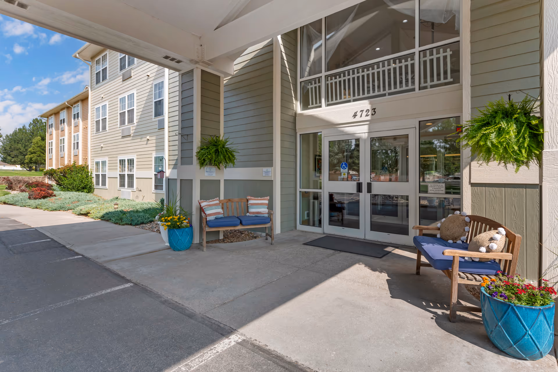 Entrance of a senior living facility with double glass doors under a covered porch. Two wooden benches with cushions and pillows are placed on either side of the entrance, accompanied by large blue flower pots with colorful flowers. The building exterior is light green with white trim, and the address number 4723 is displayed above the doors. The sky is clear and blue with some clouds.
