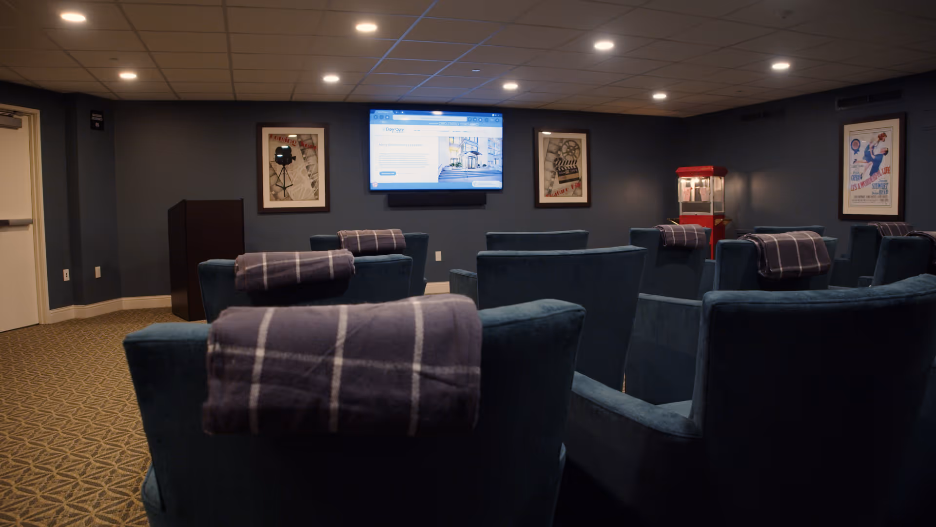 A cozy movie theater room with several blue upholstered chairs, each with a folded plaid blanket draped over the back. A large flat-screen TV is mounted on the dark blue wall at the front, displaying a webpage for Mercy Retirement and Care Center. The room is softly lit with recessed ceiling lights, and there are framed vintage movie posters on the walls. A red popcorn machine is visible in the corner.