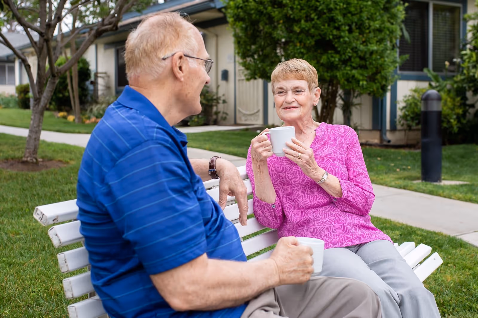 An elderly man and woman sitting on a white bench outdoors, enjoying a conversation while holding white mugs. They are surrounded by green grass, trees, and residential buildings in the background.