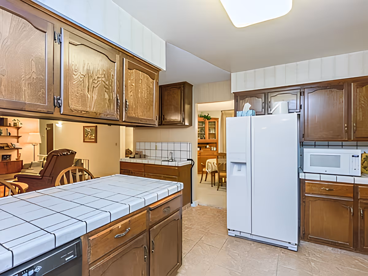 Interior view of a kitchen with wooden cabinets, a white tiled countertop, a white refrigerator, and a microwave. The kitchen opens into a dining area with a wooden table and chairs, and a living room area with a lamp and armchair is visible in the background.